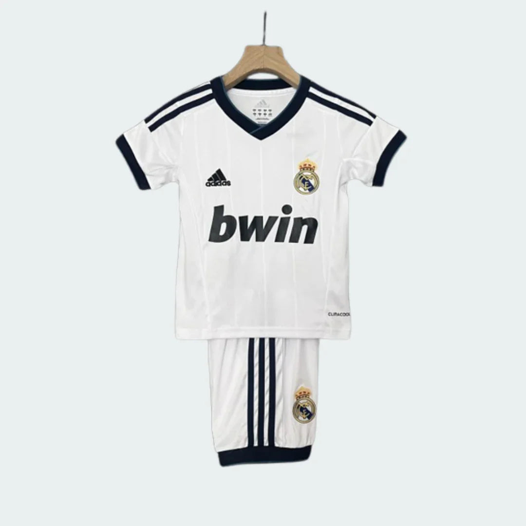 Kit infantil retrô Real Madrid I 12/13 completo, com camisa branca de gola V azul, escudo e logo Adidas. Shorts brancos com escudo.