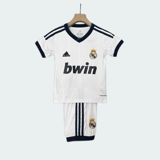 Kit infantil retrô Real Madrid I 12/13 completo, com camisa branca de gola V azul, escudo e logo Adidas. Shorts brancos com escudo.