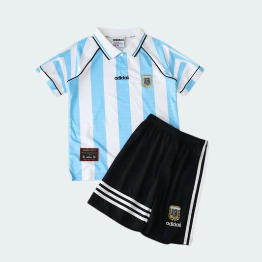 Kit infantil retrô da Argentina com camisa listrada azul celeste e branca, gola polo, e shorts pretos com listras brancas e escudos.