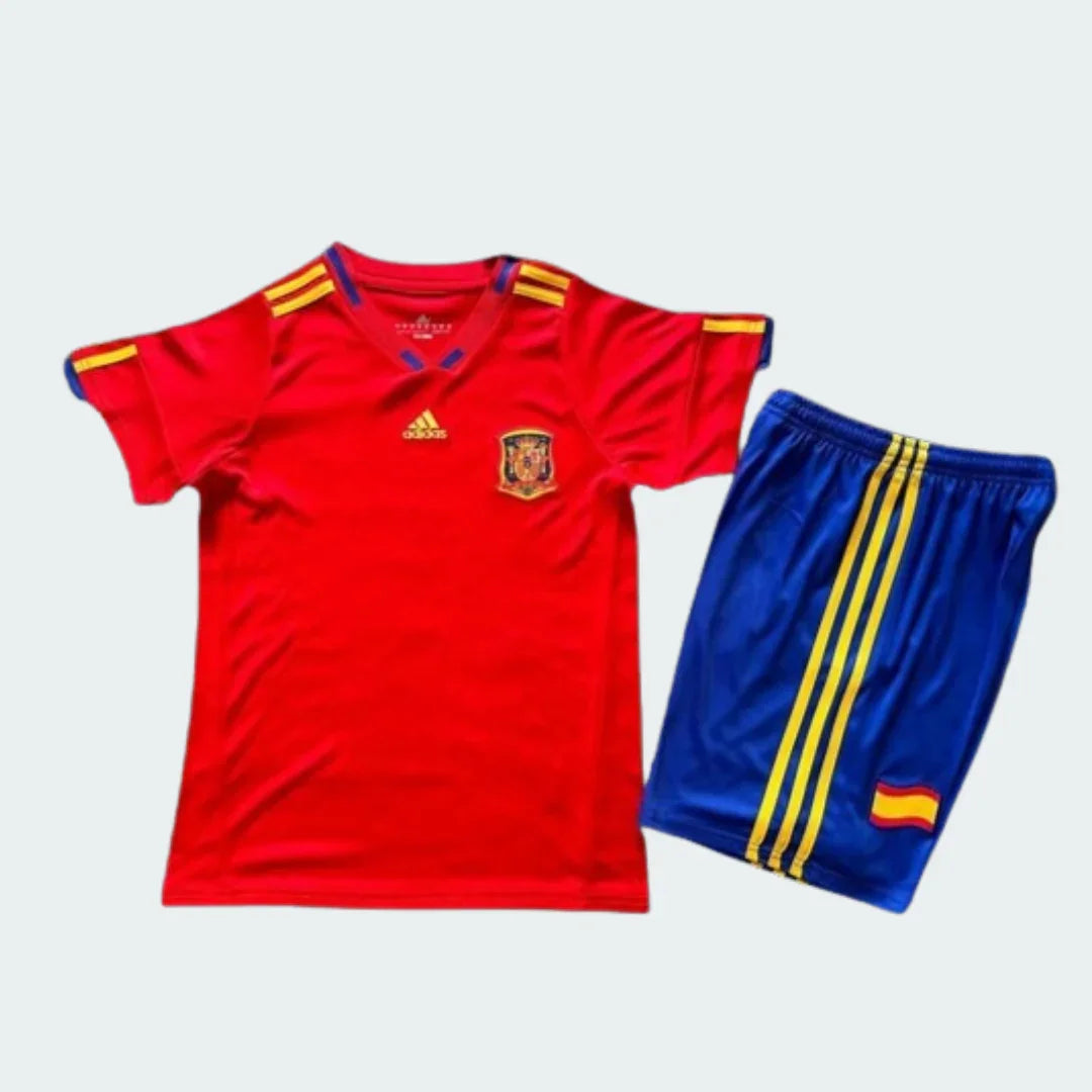 Kit infantil completo da Espanha I 2010, com camisa vermelha de mangas curtas, shorts azul, listras amarelas e escudos visíveis.