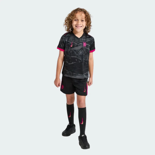 Criança sorridente veste kit de futebol do Chelsea preto com estampa cinza e detalhes rosa, shorts, meias e tênis pretos. Escudo e swoosh visíveis.
