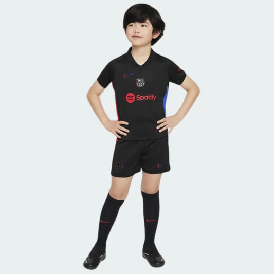 Criança em pé, vestindo kit de futebol preto do Barcelona com camisa, shorts e meias. Detalhes vermelhos, azuis, logo Spotify e escudo do time.