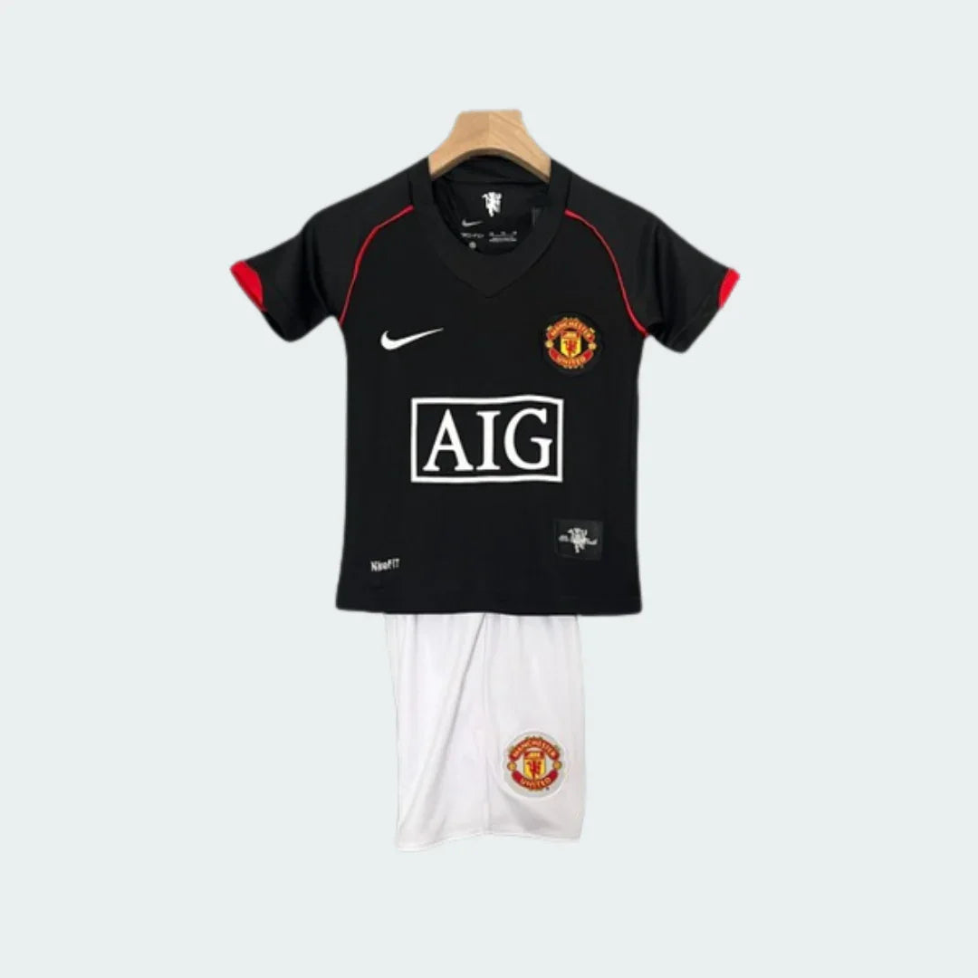 Kit infantil de futebol Manchester United preto com shorts brancos, escudo e logo AIG visíveis. Pendurado em fundo cinza.