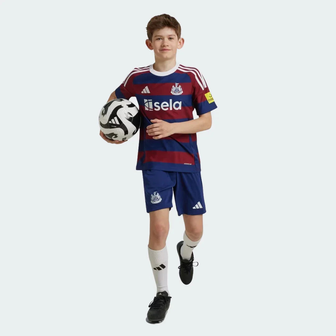 Menino sorridente em kit infantil Newcastle listrado azul-marinho e vinho, shorts azuis, meias brancas, segurando uma bola.