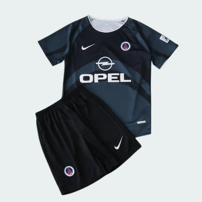 Kit infantil retrô PSG 2001. Camisa cinza escuro com faixas cinza claro, escudo PSG e Opel. Shorts pretos com escudo PSG.