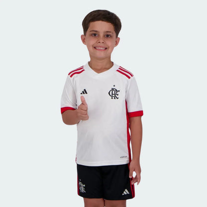 Menino sorridente vestindo kit infantil do Mengão branco e vermelho, camisa com logo Adidas e escudo CRF, shorts pretos.