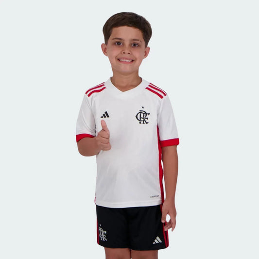 Menino sorridente vestindo kit infantil do Mengão branco e vermelho, camisa com logo Adidas e escudo CRF, shorts pretos.