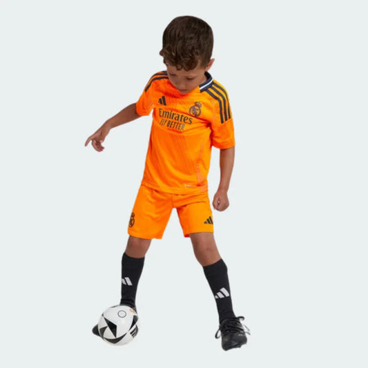 Criança em kit de futebol laranja do Real Madrid 24/25, com listras escuras nos ombros, jogando bola. Adidas e Emirates visíveis.