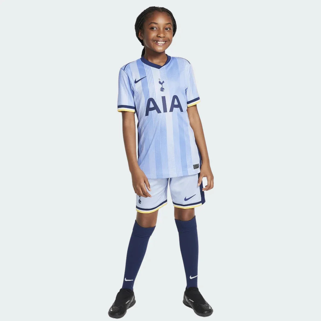 Criança sorridente veste o Kit Infantil Tottenham II 24/25, com camisa azul clara listrada, shorts e meias azul escuro. Fundo branco.