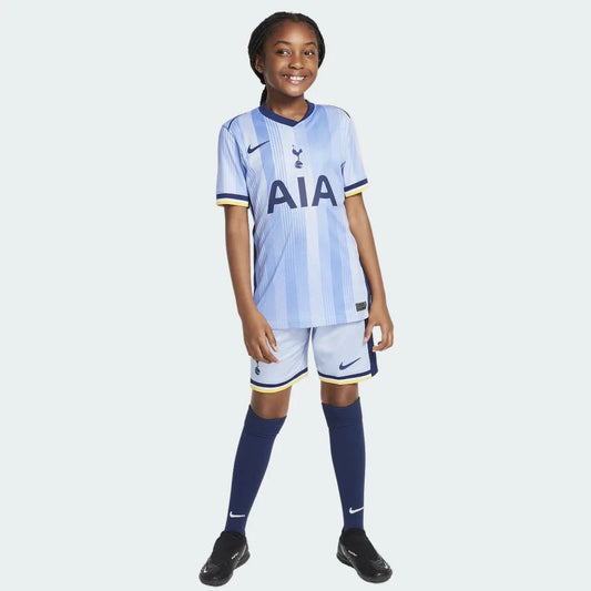 Criança sorridente veste o Kit Infantil Tottenham II 24/25, com camisa azul clara listrada, shorts e meias azul escuro. Fundo branco.