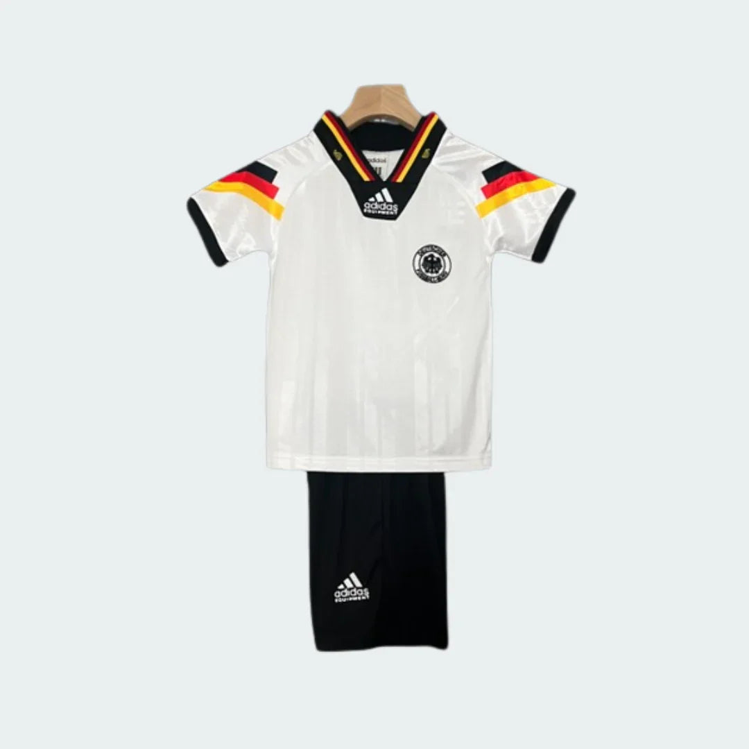 Kit infantil retrô Alemanha I 1992, com camisa branca, gola V tricolor, listras preto, vermelho e amarelo nos ombros, e shorts pretos.