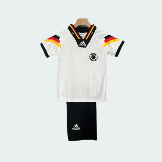 Kit infantil retrô Alemanha I 1992, com camisa branca, gola V tricolor, listras preto, vermelho e amarelo nos ombros, e shorts pretos.