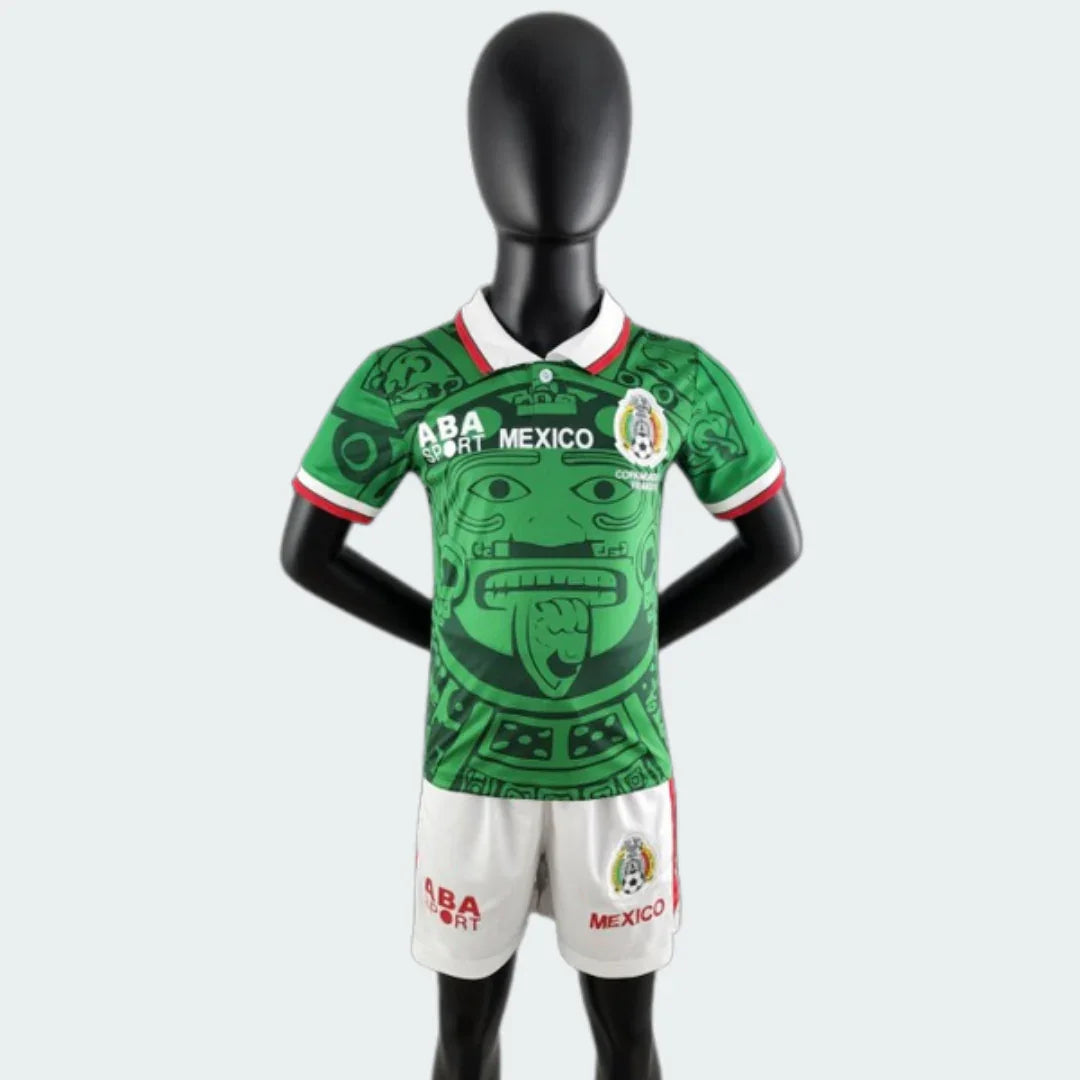 Mannequin veste kit infantil retrô Mexico. Camisa verde com estampa de rosto estilizado, gola branca. Shorts brancos.