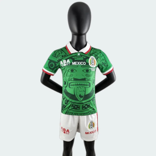 Mannequin veste kit infantil retrô Mexico. Camisa verde com estampa de rosto estilizado, gola branca. Shorts brancos.