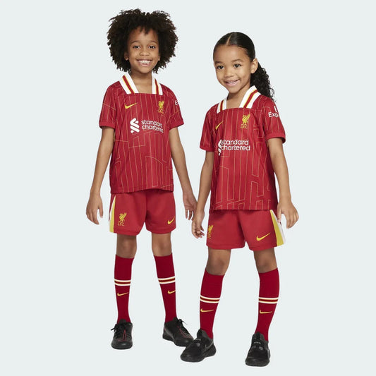Dois crianças sorrindo vestem o kit infantil vermelho do Liverpool com listras douradas, shorts vermelhos e meiões vermelhos.