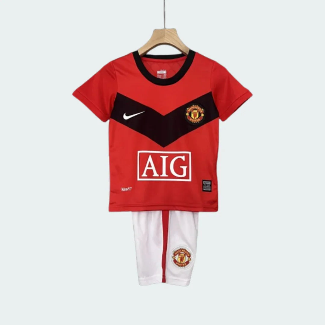 Kit infantil do Manchester United I 09/10, camisa vermelha com V preto, shorts brancos com listra vermelha e escudo do time.