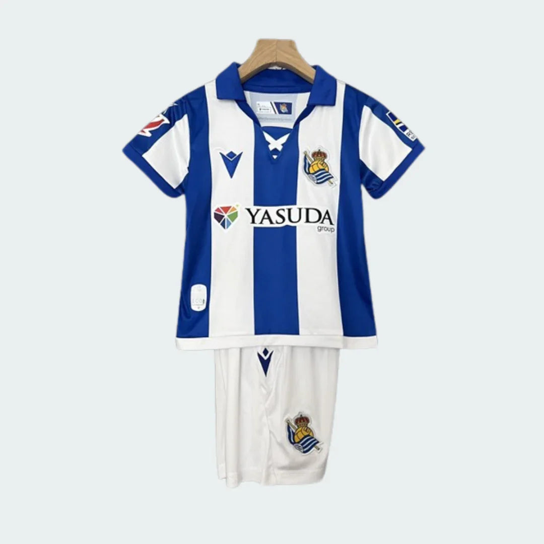 Kit infantil Real Sociedad 24/25 com camisa listrada azul e branca, gola azul, escudo do time e patrocinadores. Shorts brancos com escudo.
