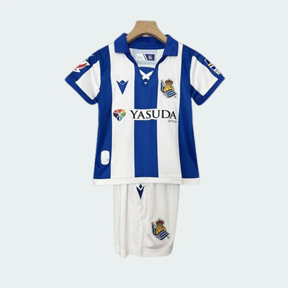 Kit infantil Real Sociedad 24/25 com camisa listrada azul e branca, gola azul, escudo do time e patrocinadores. Shorts brancos com escudo.