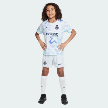Criança sorridente veste kit de futebol Inter de Milão II branco com estampas azuis, shorts brancos e meias brancas, em estúdio.