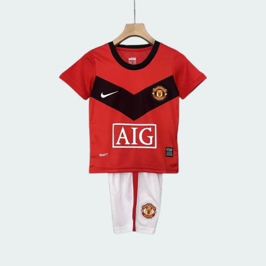 Kit infantil completo do Manchester United I 98/99, com camisa vermelha e detalhes pretos, e shorts brancos, pendurado.