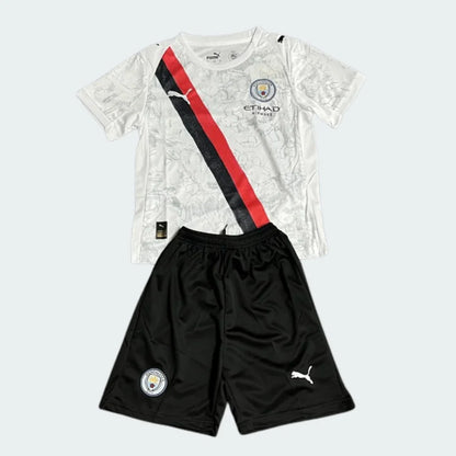 Kit infantil Manchester City Superkids 25/26. Camisa branca com faixa diagonal preta e vermelha, shorts pretos. Escudo e logos visíveis.