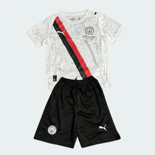 Kit infantil Manchester City Superkids 25/26. Camisa branca com faixa diagonal preta e vermelha, shorts pretos. Escudo e logos visíveis.