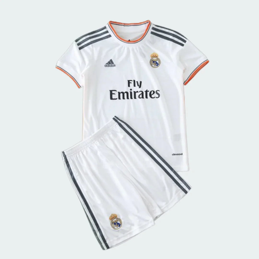 Kit infantil branco do Real Madrid com camisa e shorts. Camisa com listras cinzas nos ombros, escudo, Adidas e Fly Emirates. Shorts com listras e escudo.