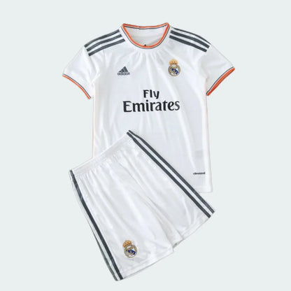 Kit infantil branco do Real Madrid com camisa e shorts. Camisa com listras cinzas nos ombros, escudo, Adidas e Fly Emirates. Shorts com listras e escudo.