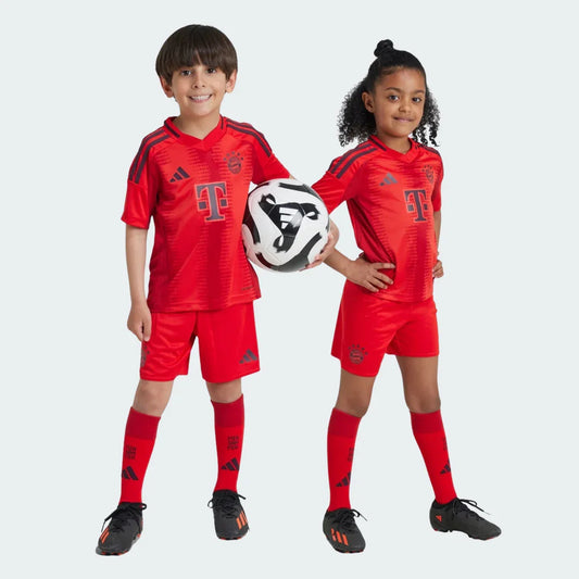 Duas crianças em kits de futebol Bayern de Munique I 24/25 vermelhos e cinza escuro. Camisa, shorts e meiões, menino com bola. Fundo branco.