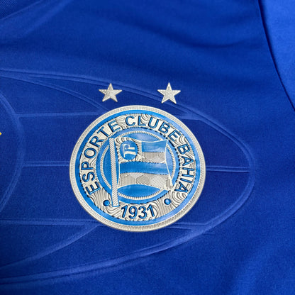 Close do escudo oficial do Esporte Clube Bahia com suas duas estrelas prateadas, aplicado sobre o tecido azul com grafismos circulares tonais.