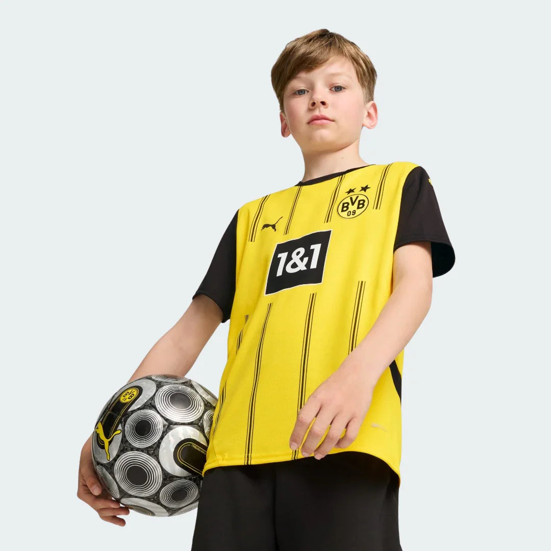 Menino com camisa amarela e preta do Borussia Dortmund, listras pretas, escudo BVB, logo Puma, patrocinador 1&1, segurando bola de futebol. Fundo branco.