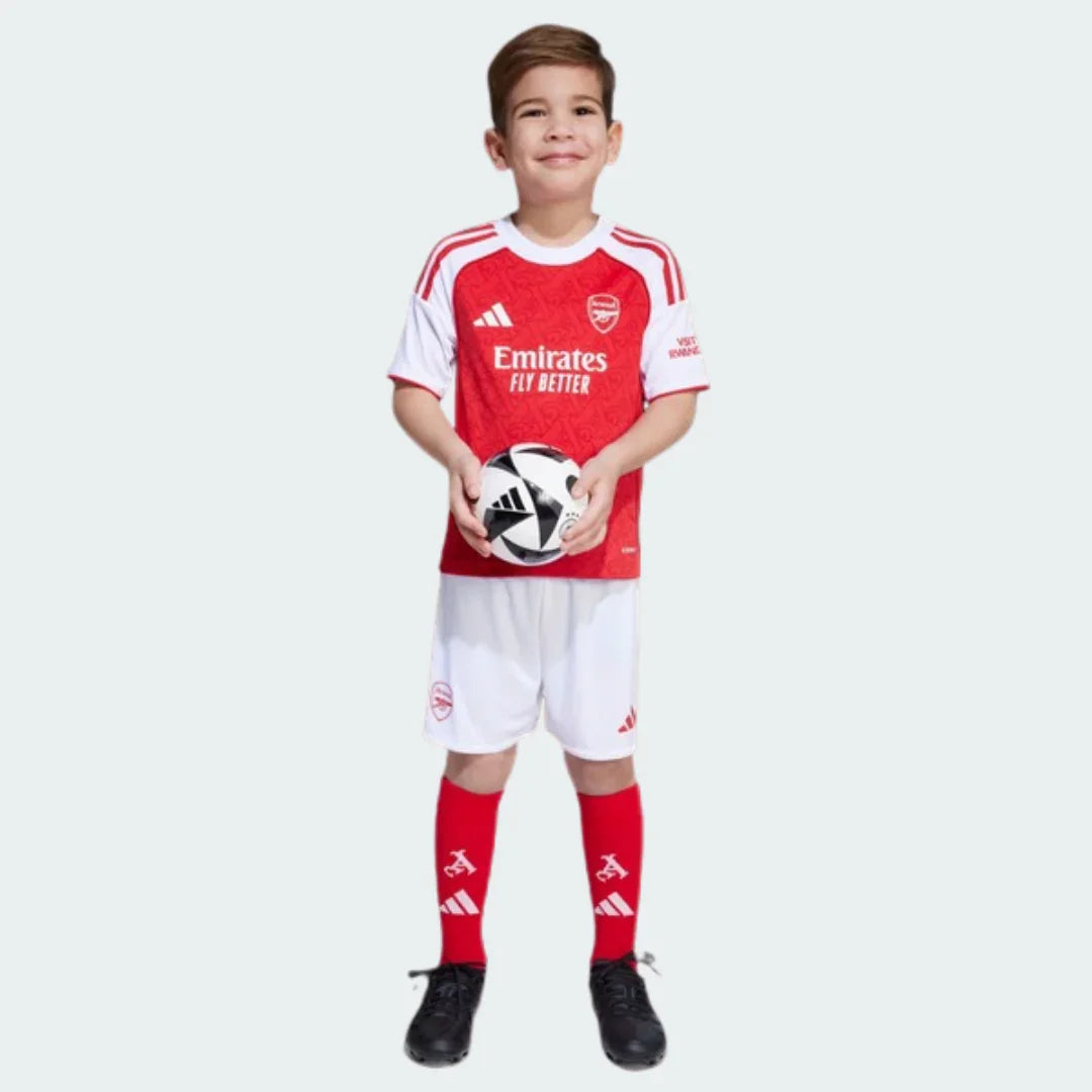 Menino sorridente em pé, vestindo kit infantil Arsenal I 25/26 vermelho e branco, segurando bola de futebol.