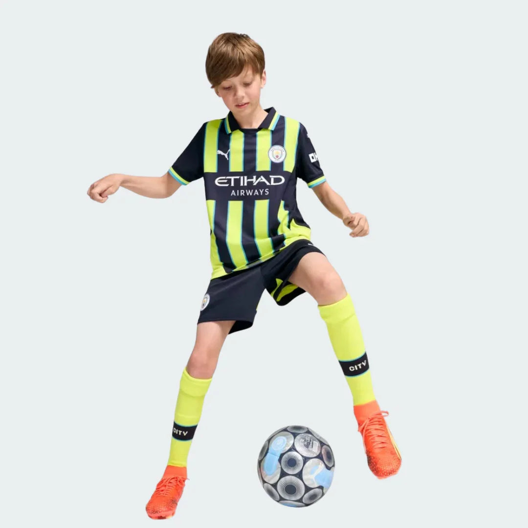 Menino usando kit de futebol Manchester City listrado em azul marinho e verde limão, shorts azuis e meias verdes, chutando uma bola.
