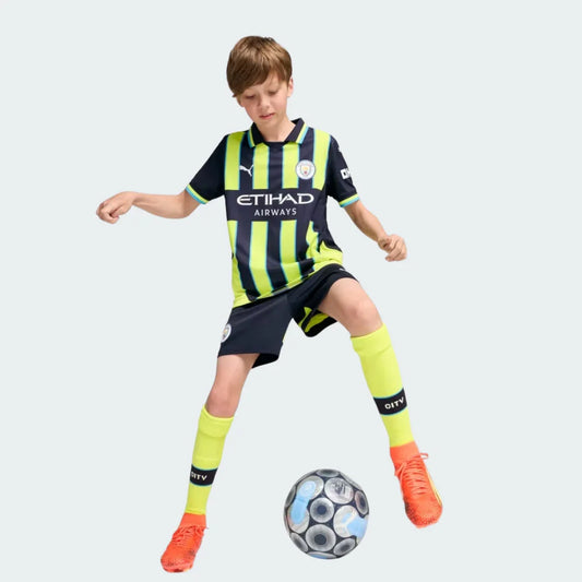 Menino usando kit de futebol Manchester City listrado em azul marinho e verde limão, shorts azuis e meias verdes, chutando uma bola.