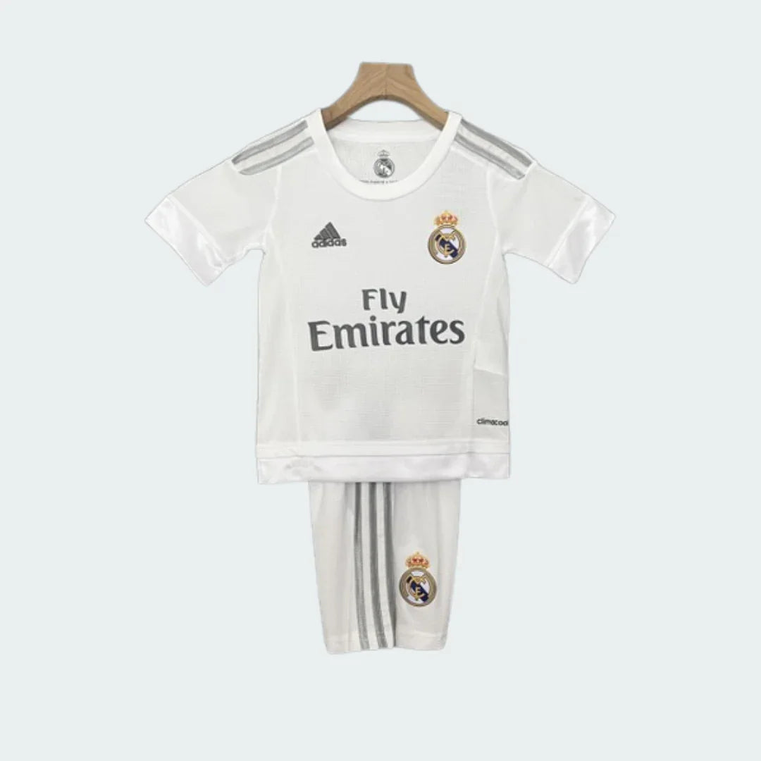 Foto frontal de um kit infantil Real Madrid I 15/16, branco com detalhes cinzas, escudo do time e logo Fly Emirates.
