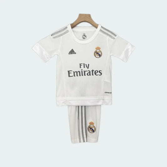 Foto frontal de um kit infantil Real Madrid I 15/16, branco com detalhes cinzas, escudo do time e logo Fly Emirates.