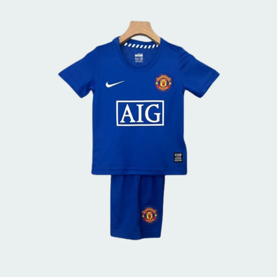 Kit infantil do Manchester United 09/10, camiseta e shorts azuis com escudo do time, logo AIG e Nike. Pendurado em cabide.