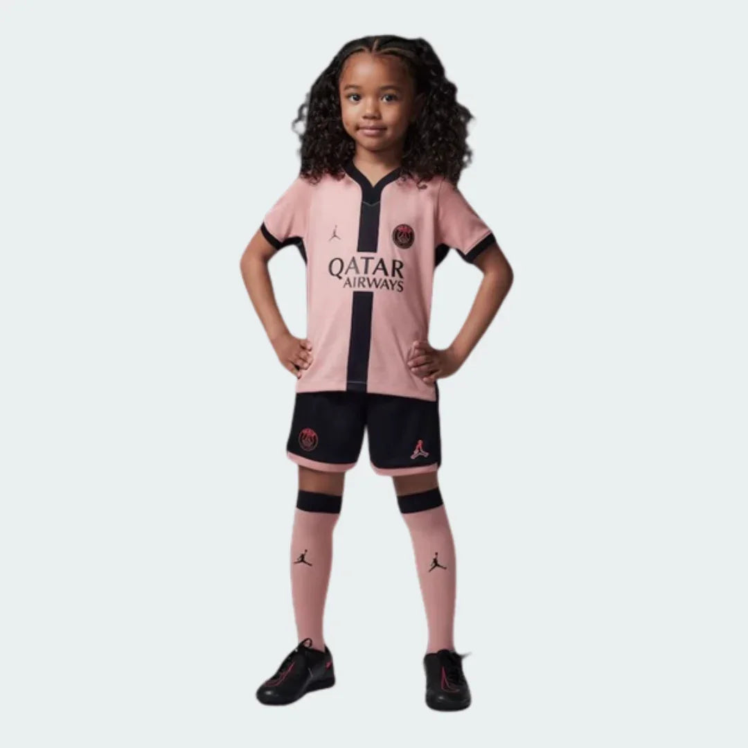 Criança sorridente vestindo kit infantil PSG 24/25. Camisa rosa com faixa preta, calção preto, meias rosa e tênis pretos.