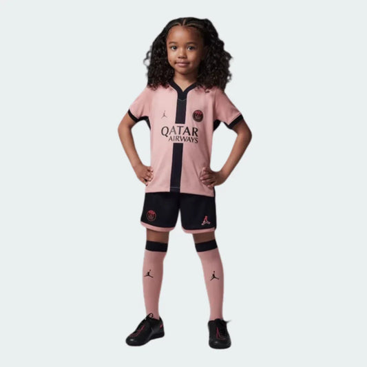 Criança sorridente vestindo kit infantil PSG 24/25. Camisa rosa com faixa preta, calção preto, meias rosa e tênis pretos.