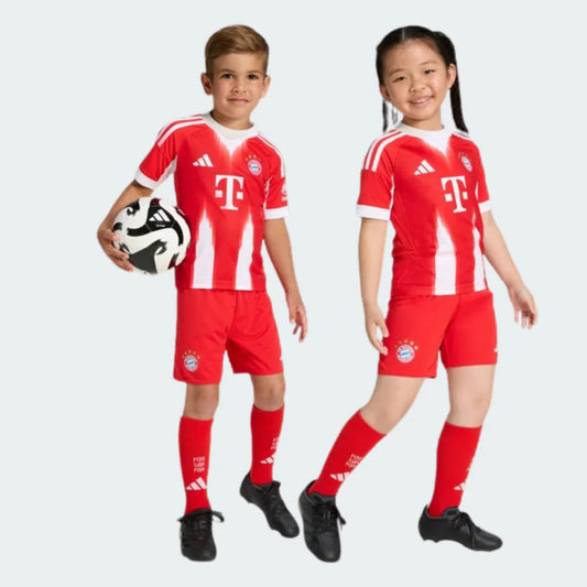 Dois jovens sorrindo com kit infantil Bayern de Munique I, camisa vermelha e branca listrada, shorts e meias vermelhas. Menino segura bola.