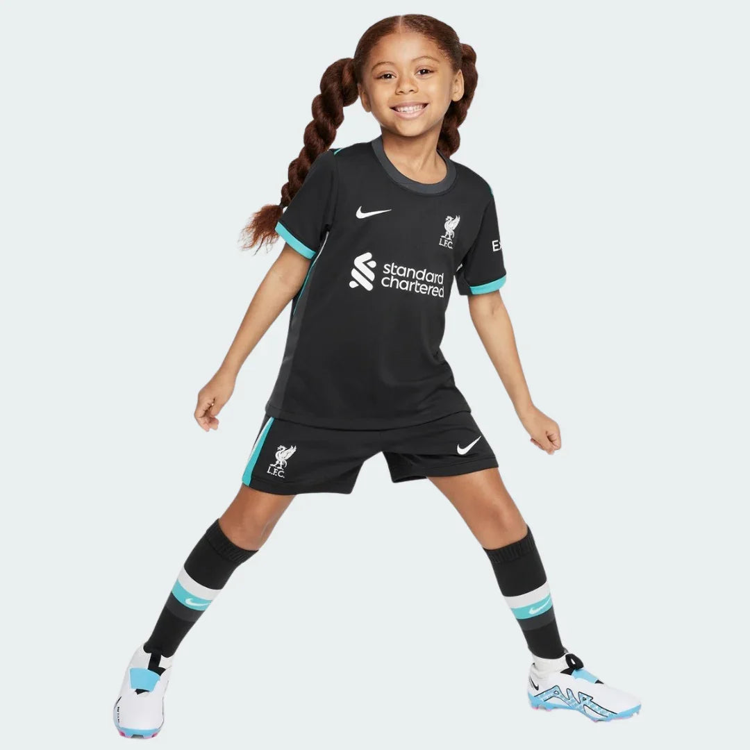 Menina sorridente vestindo kit infantil do Liverpool preto com detalhes azul claro, escudo, Nike branco e Standard Chartered.