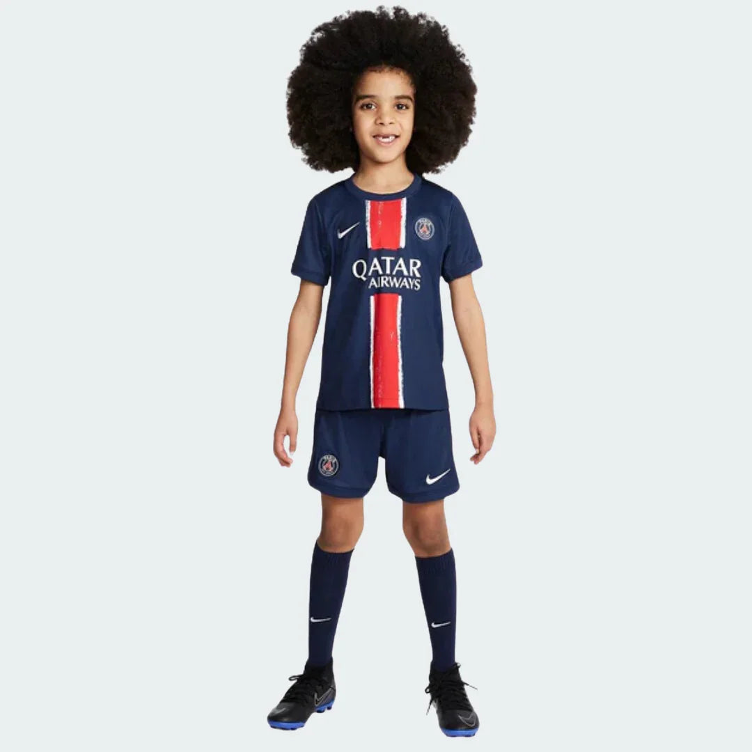 Menino sorridente em pé, vestindo o kit de futebol infantil PSG I 24/25 azul marinho com faixa vertical vermelha e shorts.