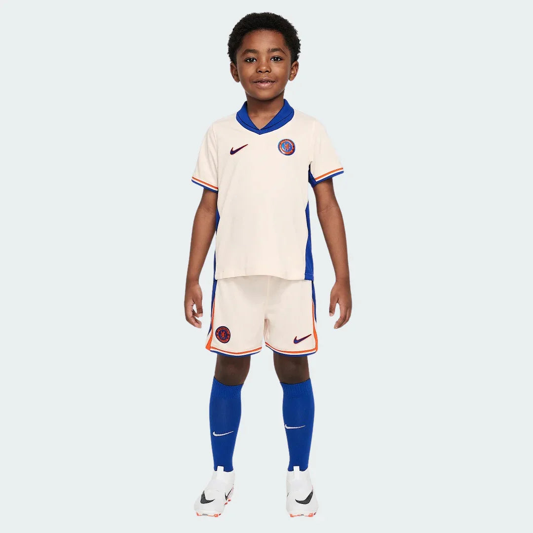 Menino sorridente veste kit infantil Chelsea 24/25 cor creme, com detalhes em azul e laranja, meias azuis e chuteiras brancas.