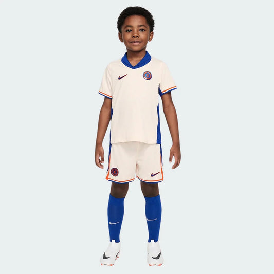 Menino sorridente veste kit infantil Chelsea 24/25 cor creme, com detalhes em azul e laranja, meias azuis e chuteiras brancas.