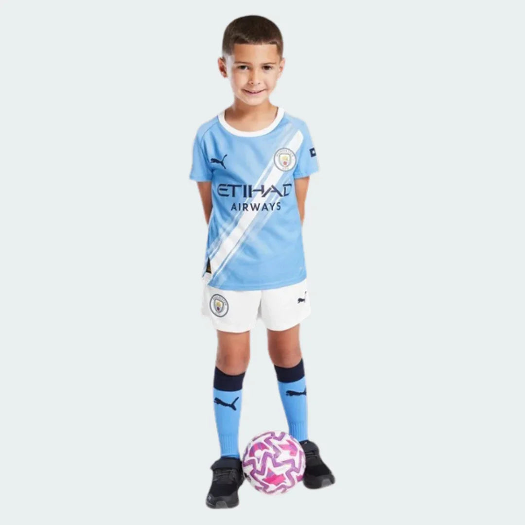 Menino sorridente veste kit infantil Manchester City azul claro, shorts branco, meias azuis, com bola de futebol rosa e branca.