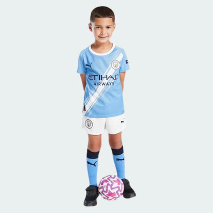 Menino sorridente veste kit infantil Manchester City azul claro, shorts branco, meias azuis, com bola de futebol rosa e branca.