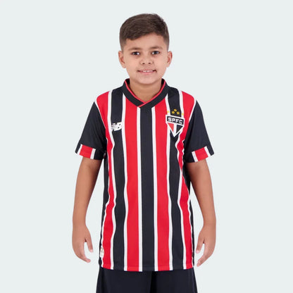 Menino sorridente em camisa infantil listrada verticalmente vermelho, preto e branco, com escudo SPFC e logo New Balance, calção preto.