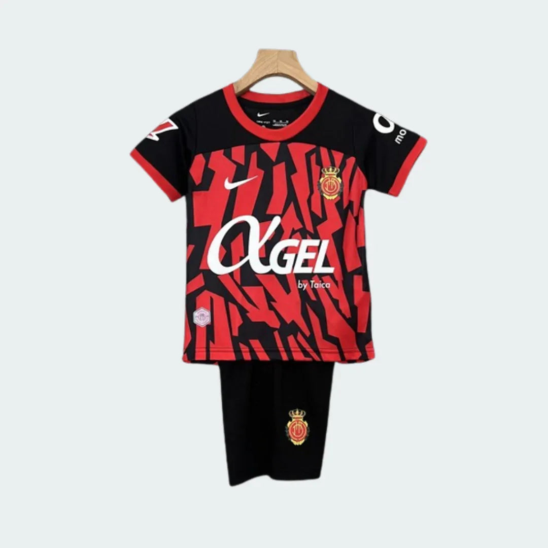 Kit infantil Mallorca 24/25, camisa preta e vermelha com padrão abstrato, shorts pretos, escudo do time e patrocinadores. Pendurado em cabide.