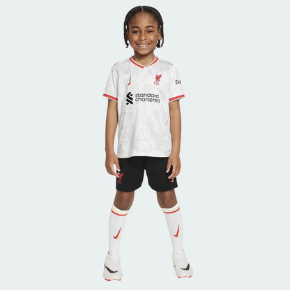 Criança sorridente veste Kit Infantil Liverpool III 24/25 completo, camisa branca com detalhes vermelhos, shorts pretos e meias brancas.