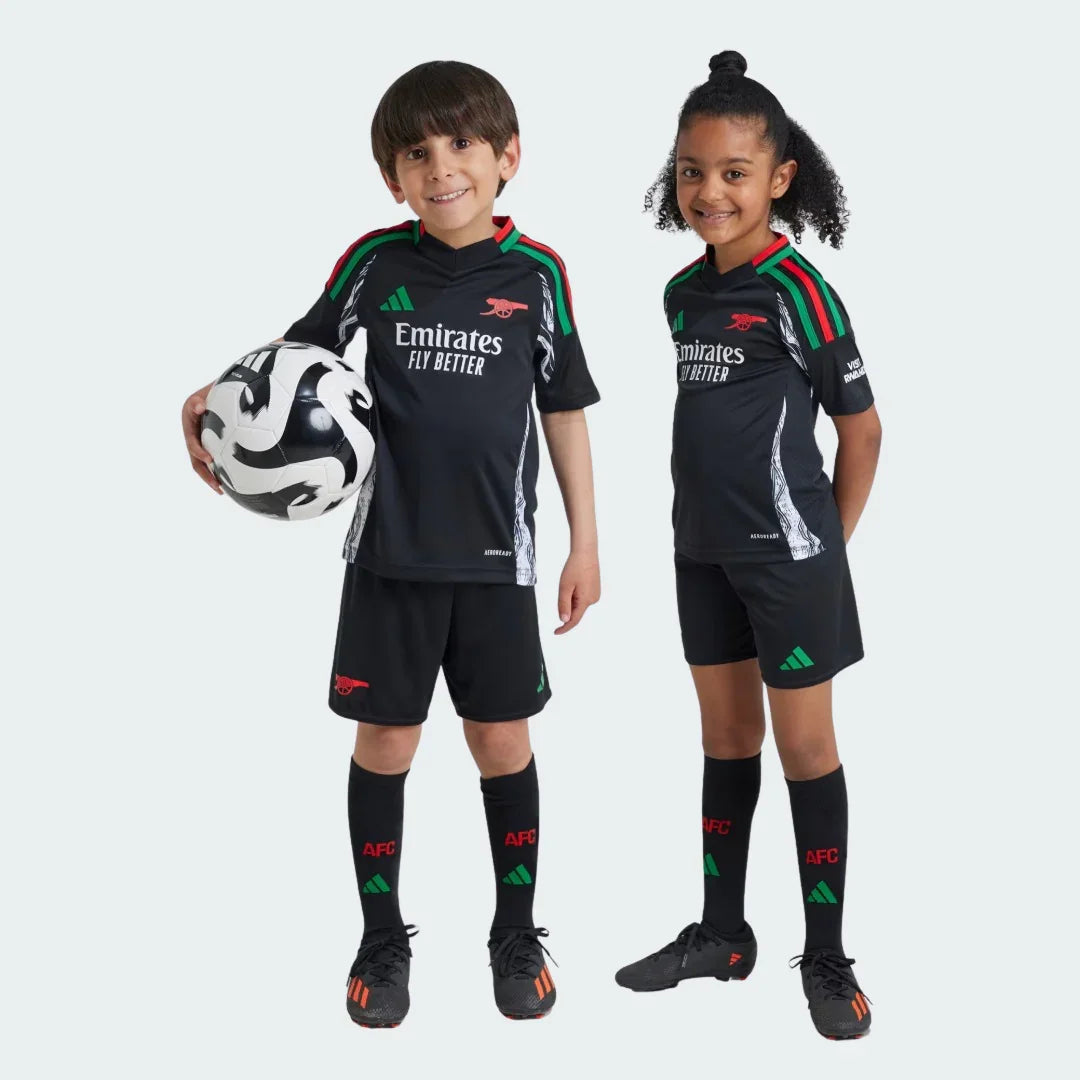 Dois jovens modelos exibem o Kit Infantil Arsenal II 24/25. A camisa é preta com detalhes verdes e vermelhos, shorts e meias pretas.